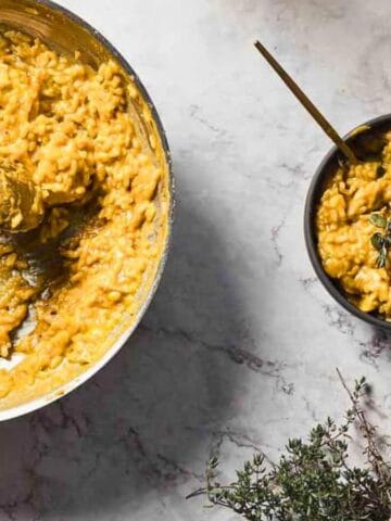 A pan filled with creamy orange-colored risotto sits on a marble countertop. A hand holds a bowl of the risotto garnished with seeds and herbs. A bundle of fresh thyme is placed nearby.