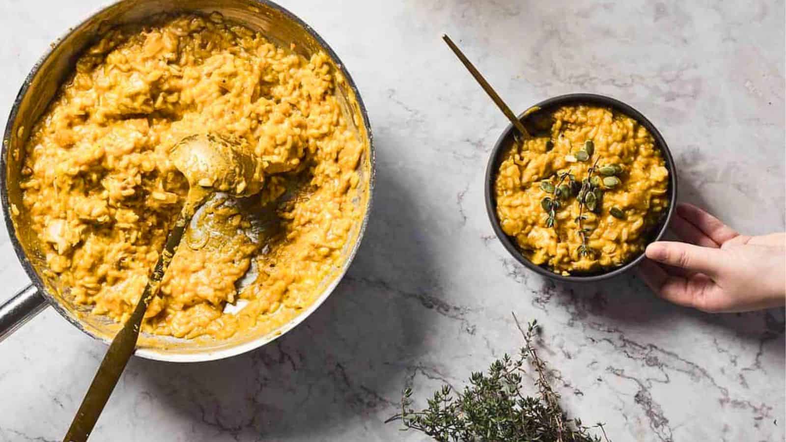 A black bowl filled with creamy pumpkin oatmeal garnished with fresh thyme and pumpkin seeds. A spoon rests inside the bowl. The background is a light gray marble surface scattered with a few pumpkin seeds.