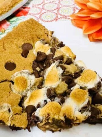 A large cookie slice topped with toasted marshmallows and chocolate chips is on a white plate. A bright orange artificial flower is next to the plate, which sits on a colorful patterned tablecloth.
