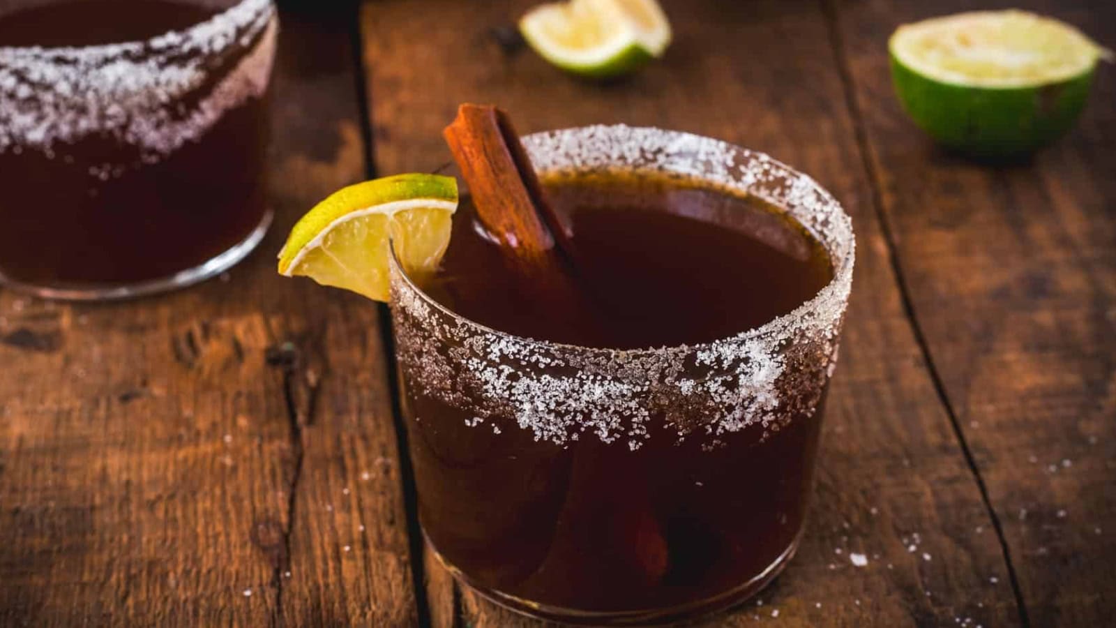 A glass filled with a dark beverage, garnished with a lime wedge and cinnamon stick, and rimmed with coarse salt. Another drink and halved limes are on a rustic wooden surface in the background.