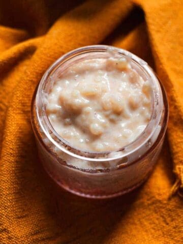 A glass jar filled with rice pudding is placed on an orange textured cloth, with two dark wooden spoons lying beside it.