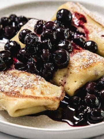 A plate with three folded crepes topped with a generous amount of blueberries in syrup and a dusting of powdered sugar.