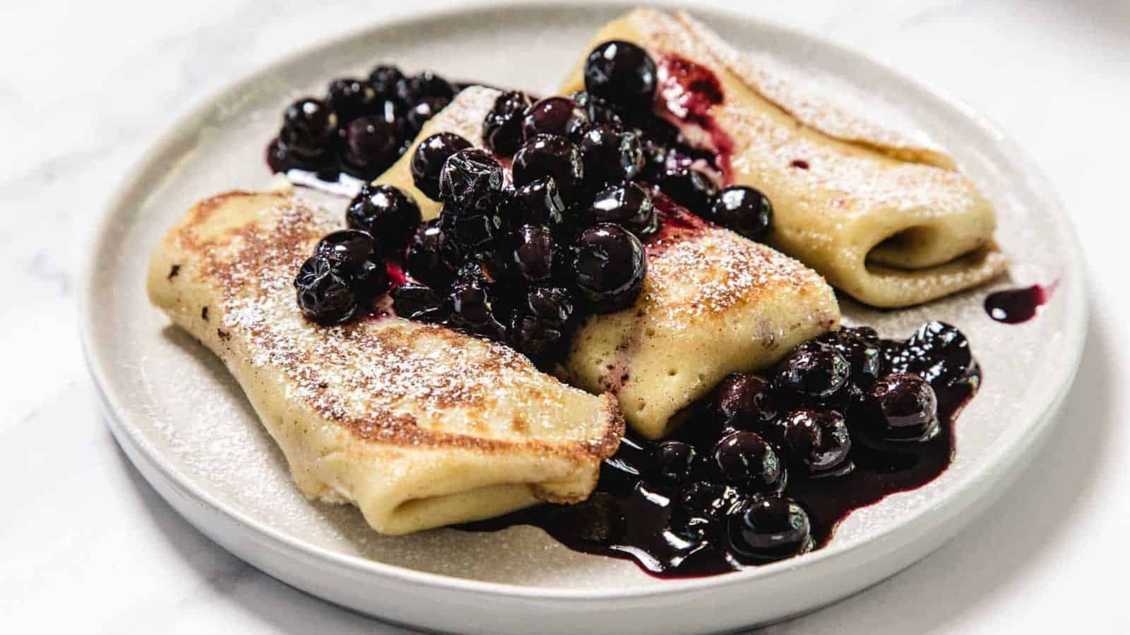 Three rolled crepes are served on a white plate, topped with powdered sugar and a generous portion of cooked blueberries in syrup. The dish sits on a light-colored surface.