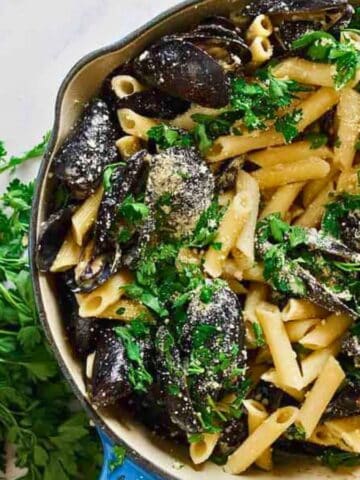 A skillet filled with penne pasta and mussels, garnished with chopped parsley and grated cheese, sits on a white marble surface with fresh parsley scattered beside it.