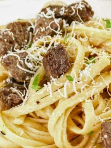 A plate of fettuccine pasta topped with pieces of beef, grated cheese, and chopped parsley, served on a white dish.