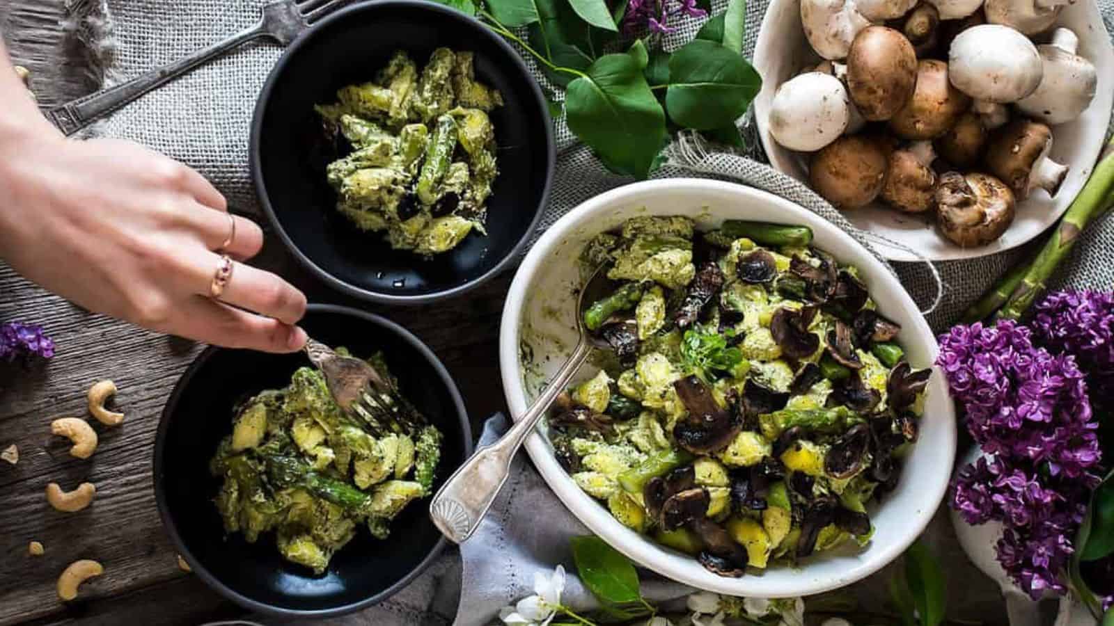 A bowl of pasta salad featuring mushrooms, asparagus, and a creamy green sauce. The dish is garnished with fresh herbs. A small bowl of nuts and some greenery are in the blurred background.