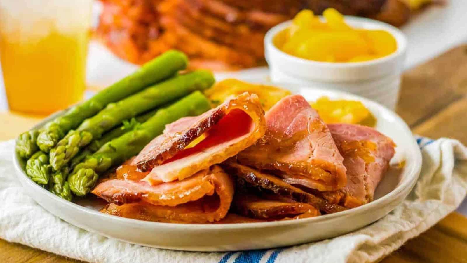 Spiral ham on a plate with asparagus and peaches in the background.