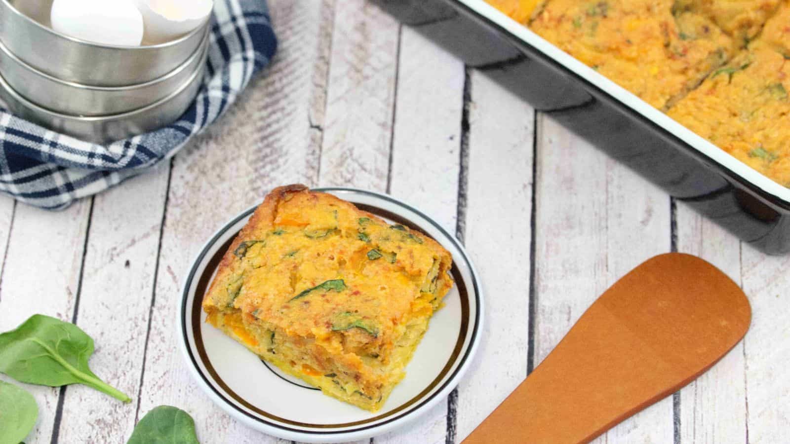 Breakfast casserole on a white plate with more casserole in a pan behind it and a blue linen with steel bowls on top beside it and wooden spoon in front of it.