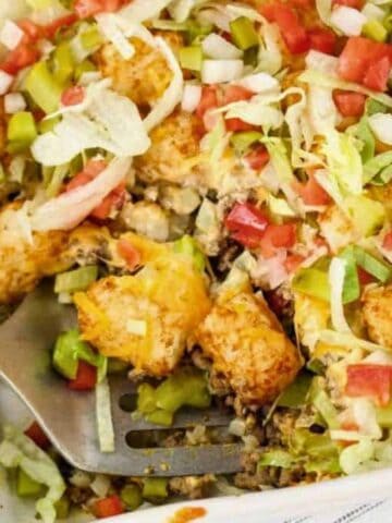 A casserole dish filled with tater tots, ground beef, shredded lettuce, diced tomatoes, pickles, and cheese, with a spatula lifting a portion from the corner.