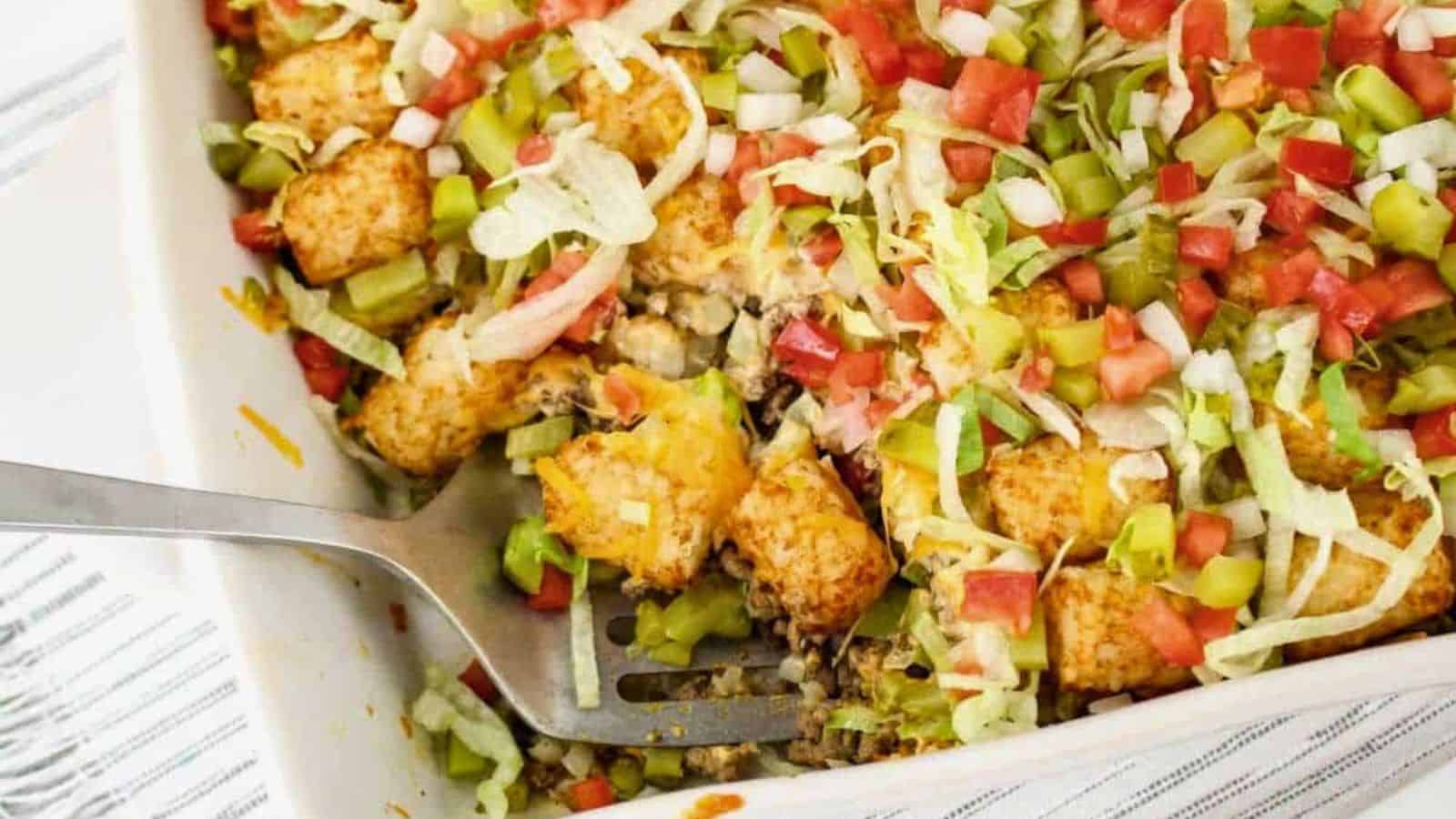 A casserole dish filled with tater tots, ground beef, diced tomatoes, shredded lettuce, onions, and pickles, with a serving spoon on the side.