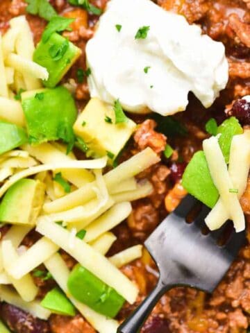 A bowl of chili topped with shredded cheese, diced avocado, and a dollop of sour cream, with a fork resting in the bowl. The food is served in a light green bowl on a green napkin.