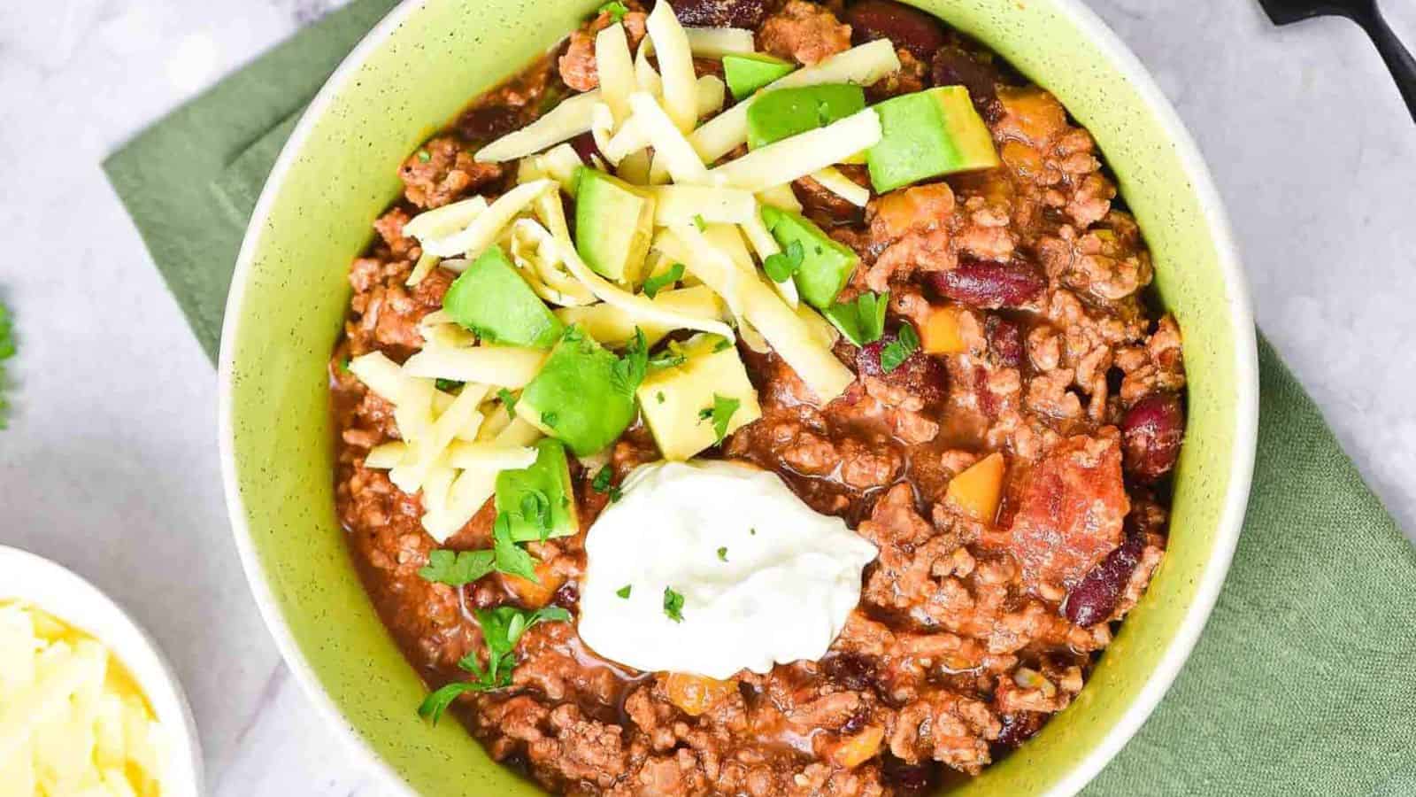 A bowl of chili topped with shredded cheese, diced avocado, and a dollop of sour cream, with a fork resting in the bowl. The food is served in a light green bowl on a green napkin.