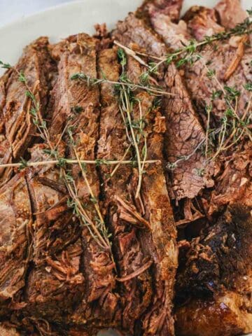 A white oval plate holds several slices of cooked, tender beef roast topped with sprigs of fresh thyme on a light marble surface.