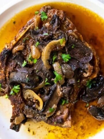A white plate containing a cooked steak topped with mushrooms, onion slices, chopped parsley, and surrounded by brown sauce.