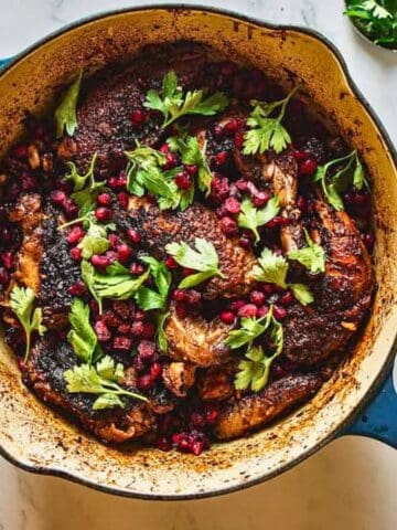 A blue Dutch oven filled with cooked chicken topped with parsley and pomegranate seeds. Surrounding it are a small bowl of walnuts, a bowl of fresh herbs, and a bowl of pomegranate seeds on a white marble surface.