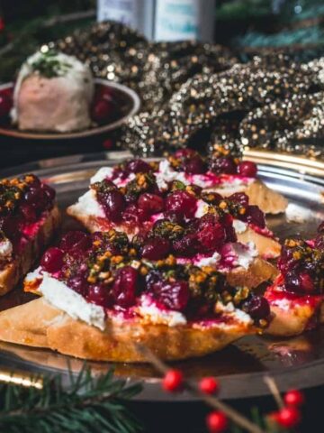 A silver tray holds slices of toasted bread topped with white spread, red berries, and chopped herbs. A small round ball covered in herbs sits in the background among festive decorations.