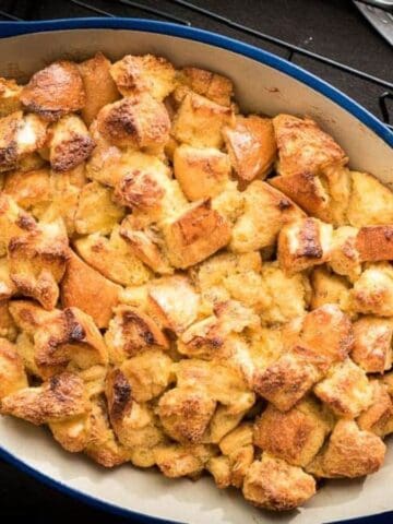 A person holds a blue oval baking dish filled with golden-brown bread pudding on a cooling rack, with utensils and a plate nearby.