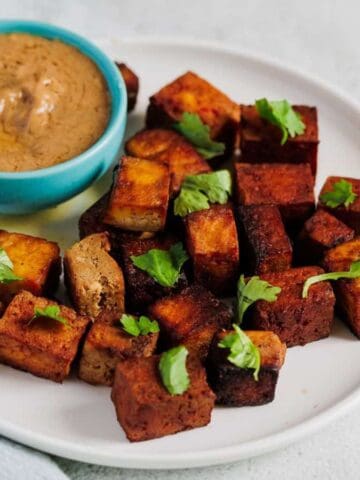 A white plate with seasoned tofu cubes garnished with cilantro, served alongside a small blue bowl of dipping sauce. The plate is set on a light surface next to a light blue napkin.