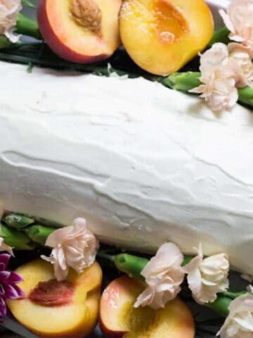 A rectangular cake covered in white frosting is surrounded by halved peaches, pink carnations, purple flowers, and green leaves on a white platter.