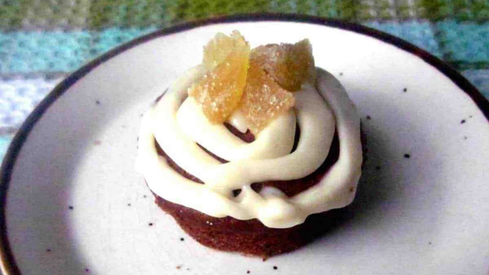 A small, round dessert sits on a white plate with a dark rim. The treat is topped with a swirl of white icing and pieces of candied ginger. A checkered cloth is visible in the background.