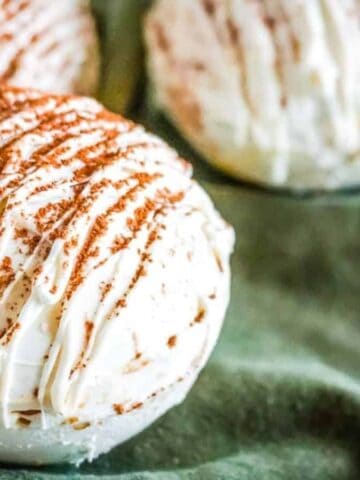 Three round white desserts sit on a dark green cloth. Each dessert is coated in white icing and drizzled with more icing, then sprinkled with a brown powder, possibly cinnamon or cocoa.