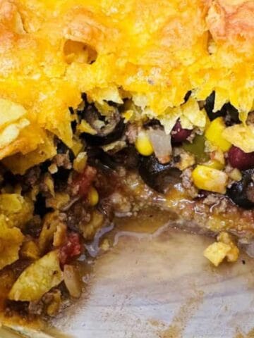 A baked casserole with a golden cheese topping is shown in a glass dish. A section has been scooped out, revealing layers of ground beef, corn, kidney beans, tomatoes, black olives, and tortilla chips inside.