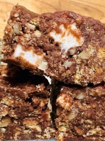 Three chocolate dessert bars with visible chunks of marshmallow and nuts are stacked on a plate. Blurred jars of chocolate chips and marshmallows are in the background.