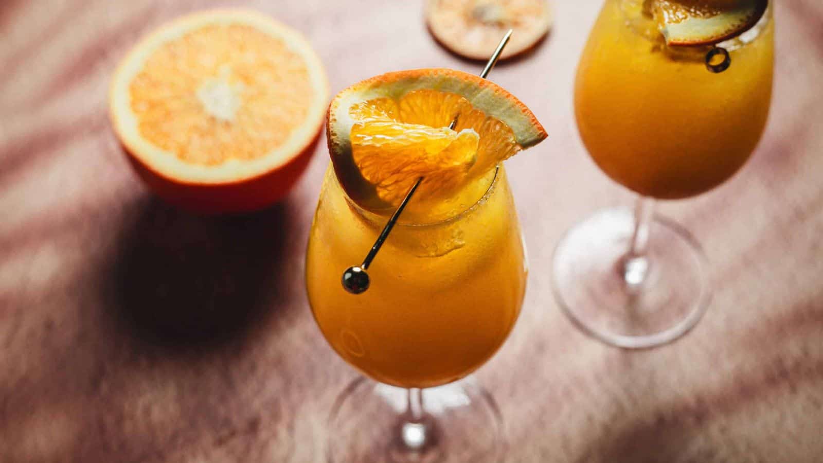 Two glasses of orange-colored cocktails garnished with orange slices and skewers are on a wooden surface. A halved orange is also visible in the background.