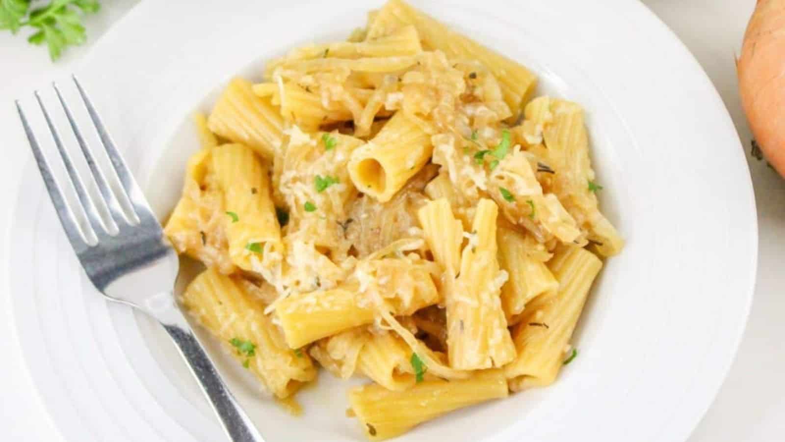 A plate of rigatoni pasta with shredded cheese and herbs, next to a fork on a white dish. An onion and parsley are in the background.