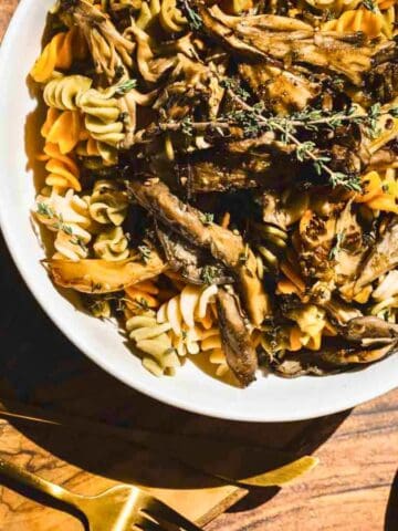 A white bowl filled with rotini pasta and topped with shredded mushrooms and fresh herbs sits on a wooden surface. A fork, knife, and small bowl of salt are placed nearby. Sunlight casts shadows over the scene.