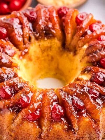 A pineapple upside-down bundt cake topped with glazed cherries sits on a white plate. Slices of pineapple and a bowl of cherries are visible in the background.