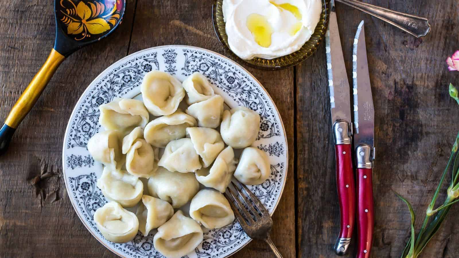 A plate of dumplings is served on a patterned dish with a fork, alongside a bowl of sour cream, a decorative spoon, and two knives with red handles on a wooden table.
