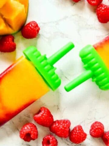 Two homemade popsicles with green handles, featuring layered yellow and red colors, are placed on a white marble surface surrounded by fresh raspberries and sliced mango pieces.