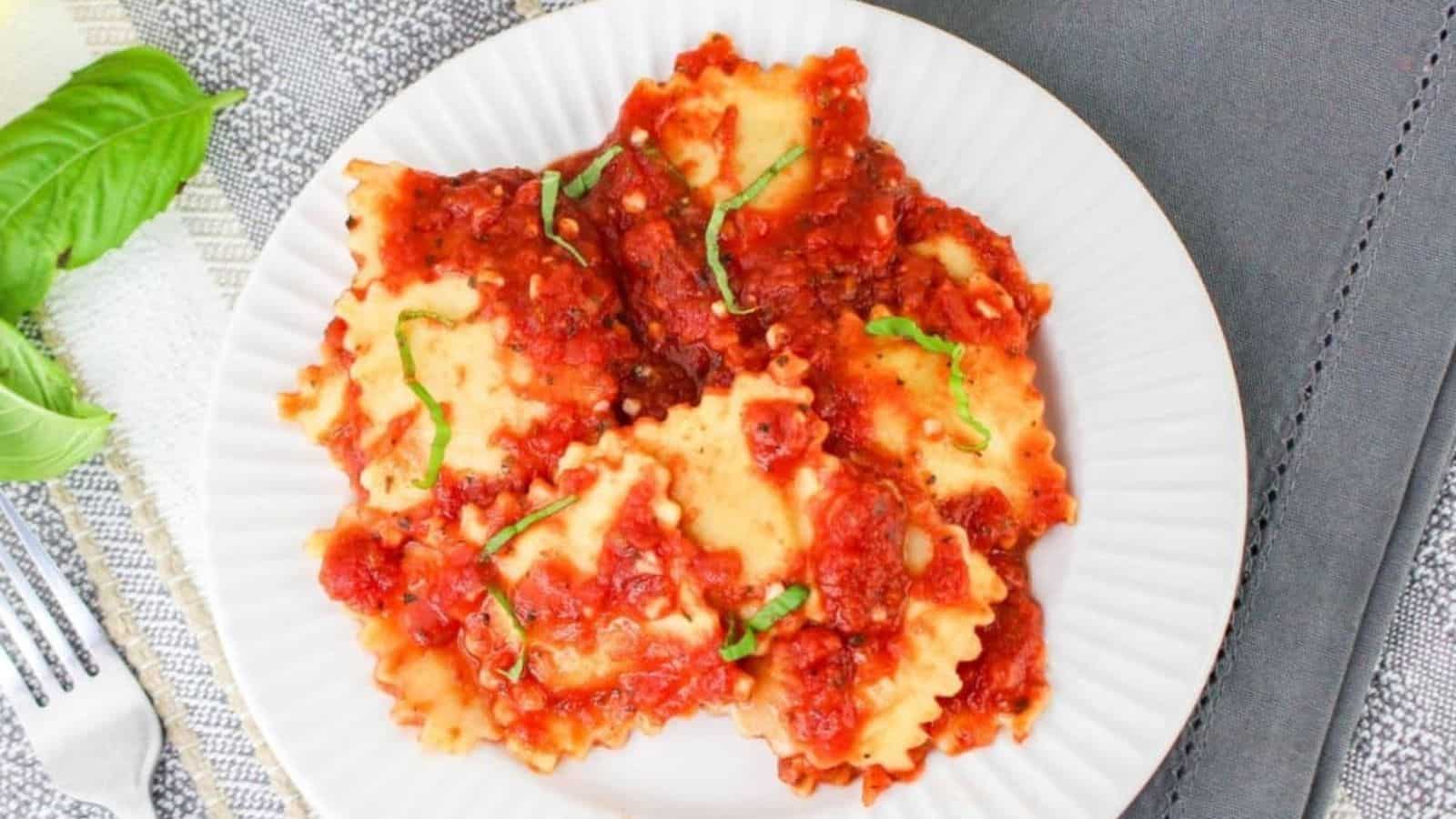 A plate of ravioli topped with red tomato sauce and garnished with fresh basil, placed on a gray tablecloth next to a fork and basil leaves.