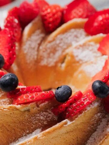 A Bundt cake topped with powdered sugar, fresh strawberries, blueberries, and raspberries sits on a white plate.