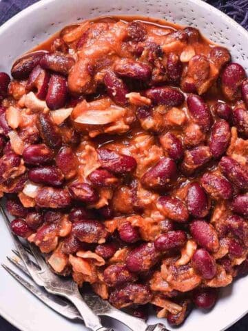 A white bowl filled with a red kidney bean stew, including visible pieces of meat or vegetables, sits on a dark cloth napkin with a fork and spoon beside it. A small bunch of parsley is on the table nearby.