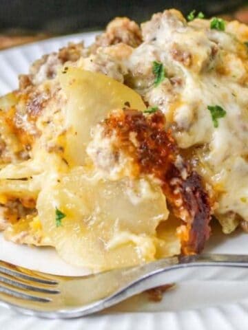 A serving of cheesy, baked ground beef and potato casserole sits on a white plate with a fork. The dish has visible layers of sliced potatoes, ground meat, and melted cheese, garnished with chopped herbs.
