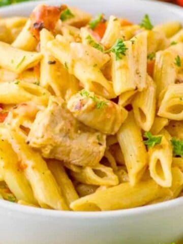 A white bowl filled with penne pasta mixed with chunks of chicken, diced red bell peppers, and garnished with chopped parsley, placed on a yellow cloth.