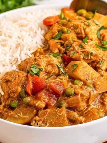 A bowl filled with white rice and a chicken curry with peas, potatoes, carrots, and sauce. A spoon rests in the bowl, and naan bread and fresh cilantro are on the side.