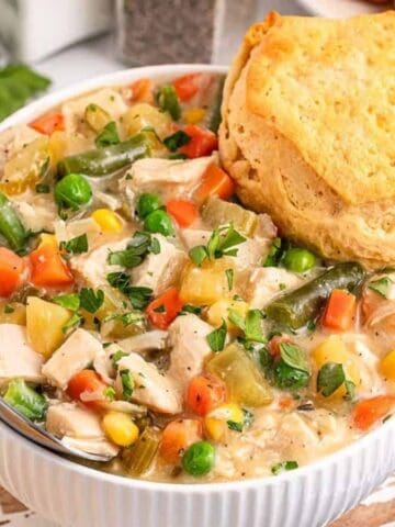 A white bowl filled with creamy chicken and vegetable stew, including carrots, peas, green beans, and corn, garnished with herbs, with a golden biscuit resting on top. A spoon is in the bowl.