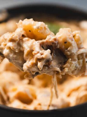 A close-up of a spoonful of creamy macaroni and cheese being lifted from a black bowl, showing melted cheese and visible pieces of pasta.
