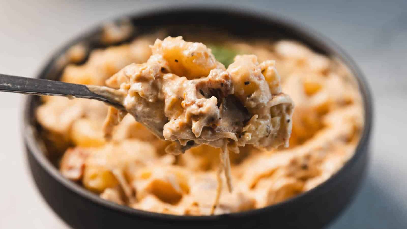 A black bowl filled with creamy pasta coated in a thick sauce, topped with a fresh basil leaf. A metal spoon rests inside the bowl. The background is a light-colored surface.