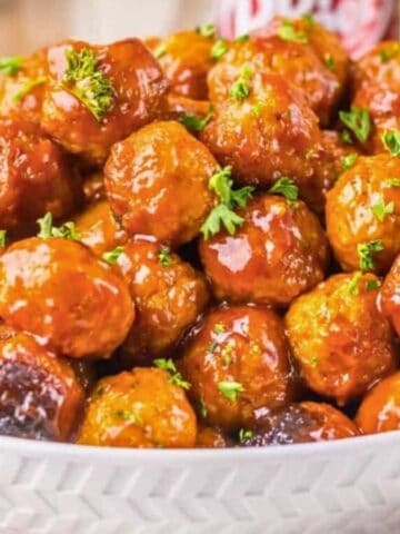A white bowl filled with glazed cocktail meatballs garnished with chopped parsley, set on a wooden surface with a white napkin on the side.
