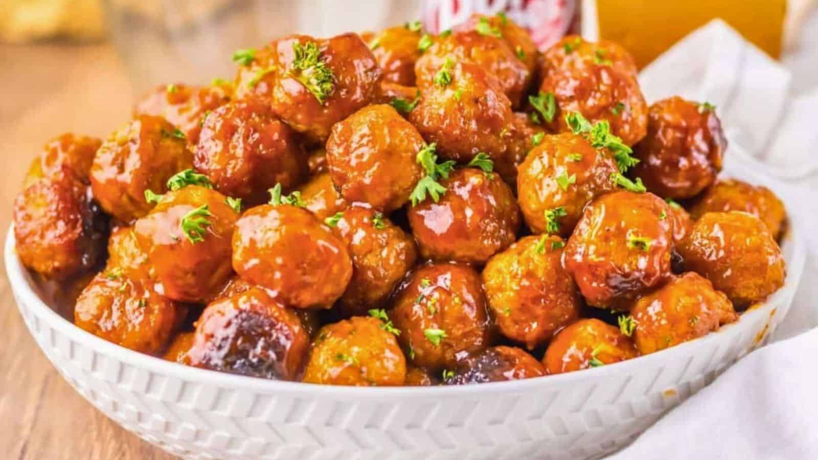 A white bowl filled with glazed meatballs, garnished with chopped parsley, placed on a wooden table with a soft drink can and a glass in the background.