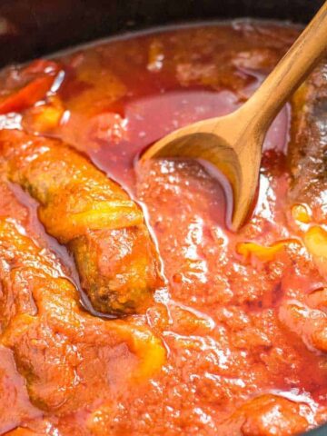 A wooden spoon rests in a pot of fish stew with a tomato-based sauce. Pieces of cooked fish are visible in the thick red sauce, along with slices of cooked peppers.