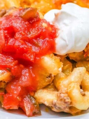 A plate of cheesy pasta casserole with ground meat, topped with red salsa and a dollop of sour cream. The dish is served on a white plate.