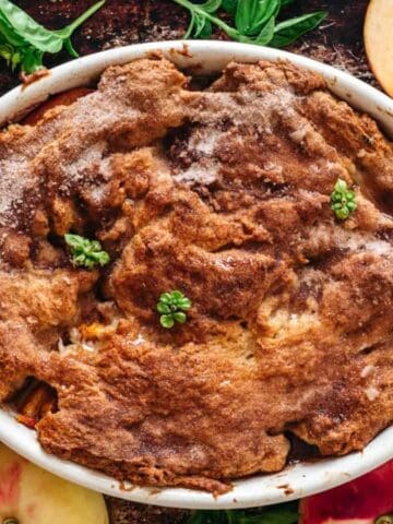 A baked peach cobbler in a white oval dish, topped with sprigs of fresh herbs, is surrounded by whole and halved peaches and green basil leaves on a dark surface.