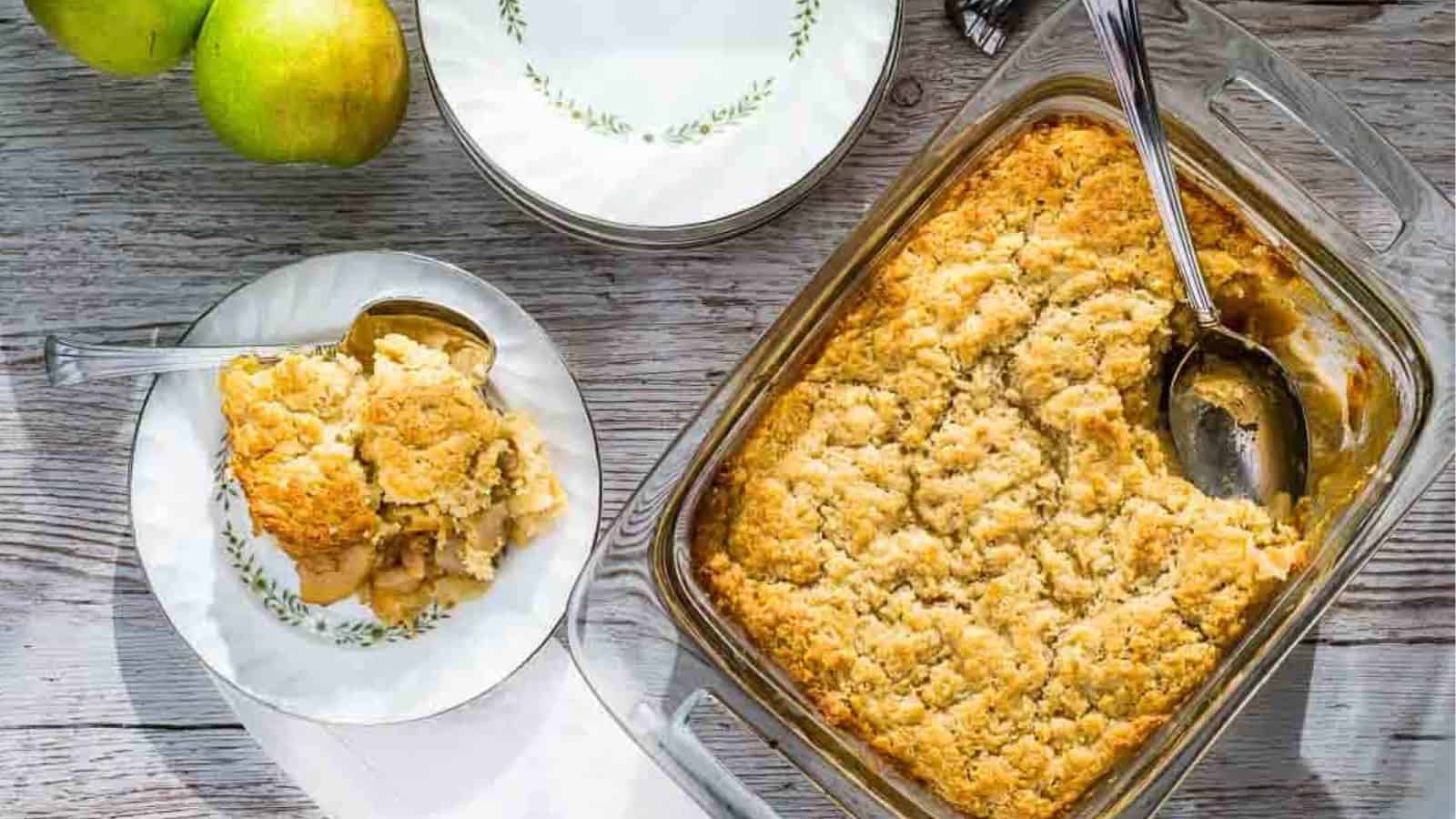 A glass baking dish with a baked apple cobbler sits on a wooden surface. Part of the cobbler is served on a small plate with a spoon. Nearby are stacked plates and two green apples.