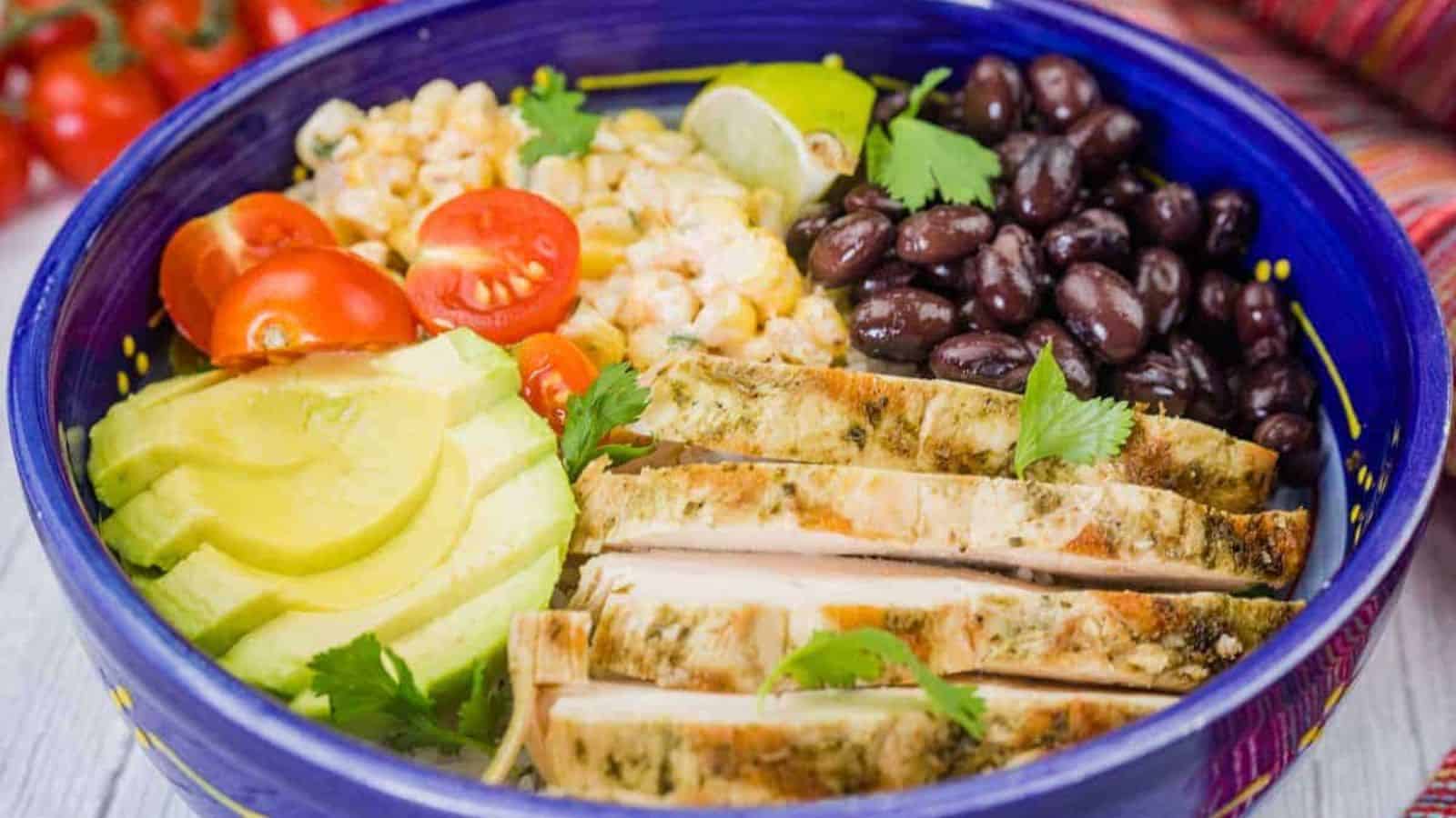 A blue bowl filled with street corn chicken rice bowl, featuring grilled chicken, corn, tomatoes, avocado, and black beans.