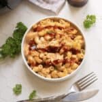 A white bowl filled with macaroni and cheese topped with barbecue sauce and chunks of chicken, placed on a white hexagonal tile surface with parsley garnish, and a fork and knife beside it.